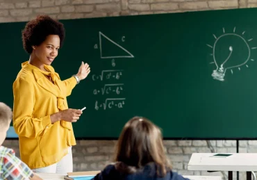 Professora ensinando matemática em frente a um quadro negro para alunos do fundamental / Concurso Prefeitura de São Carlos